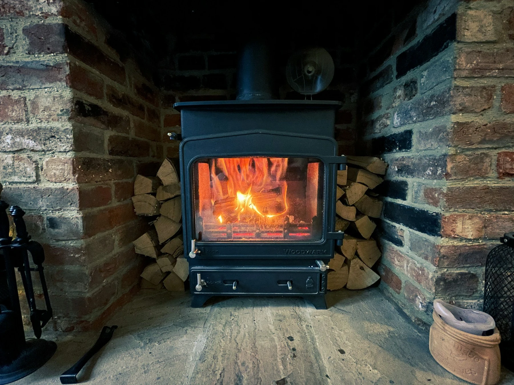 Cosy wood-burning stove with glowing flames and logs stacked beside a brick hearth